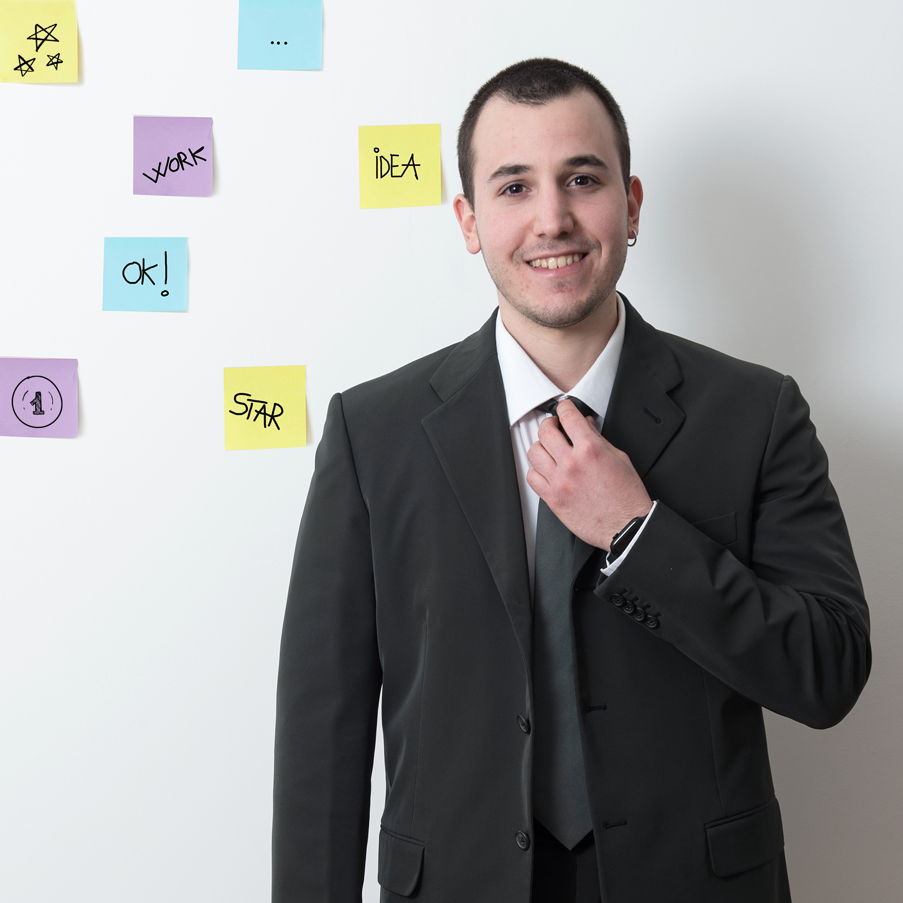 Ritratto fotografico di un giovane professionista sorridente in giacca e cravatta davanti a una parete di post-it colorati e appunti creativi