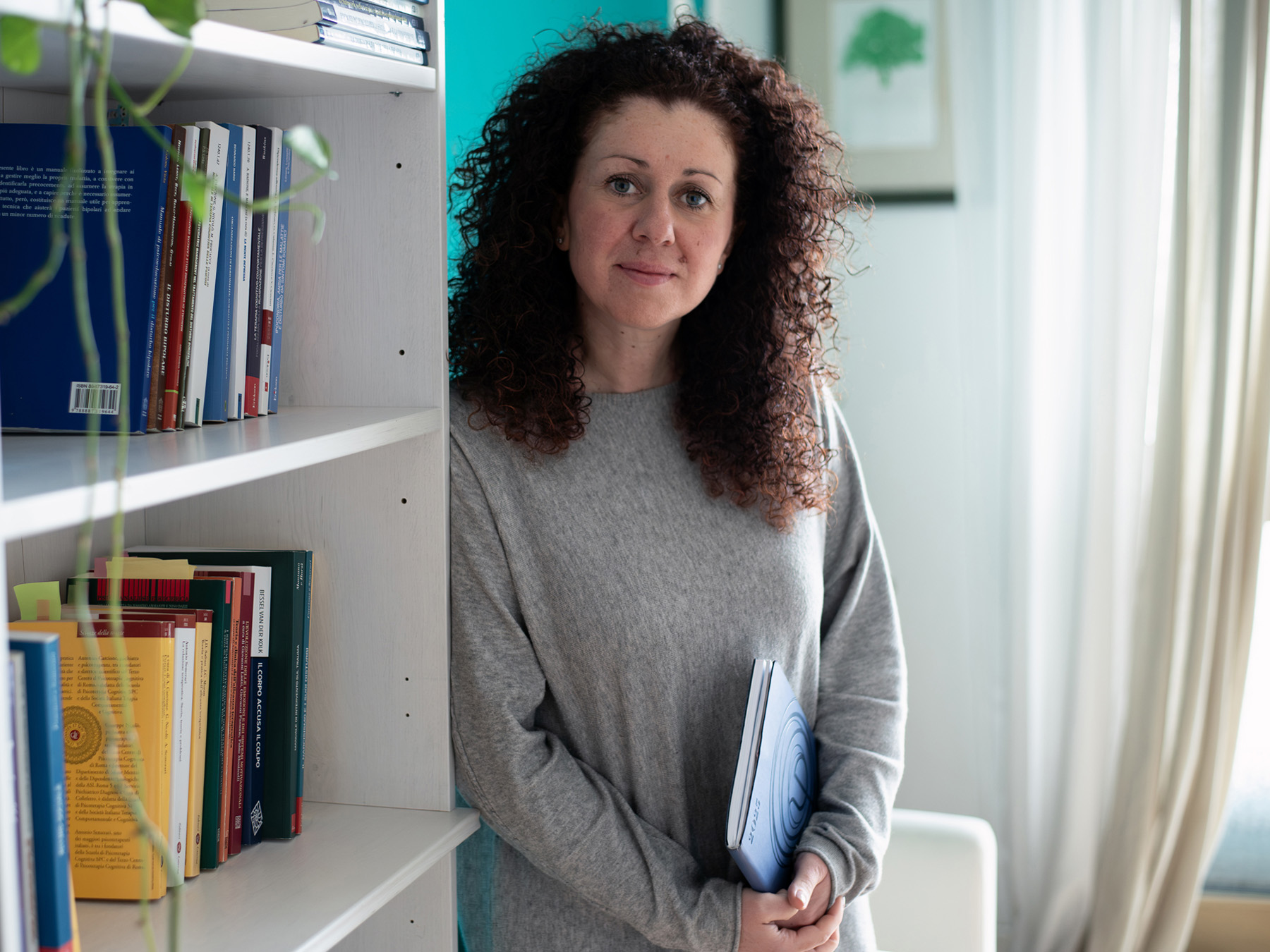 Ritratto professionale di una psicologa in piedi, appoggiata alla libreria del suo studio con una agenda in mano, con finestra e tenda nello sfondo.