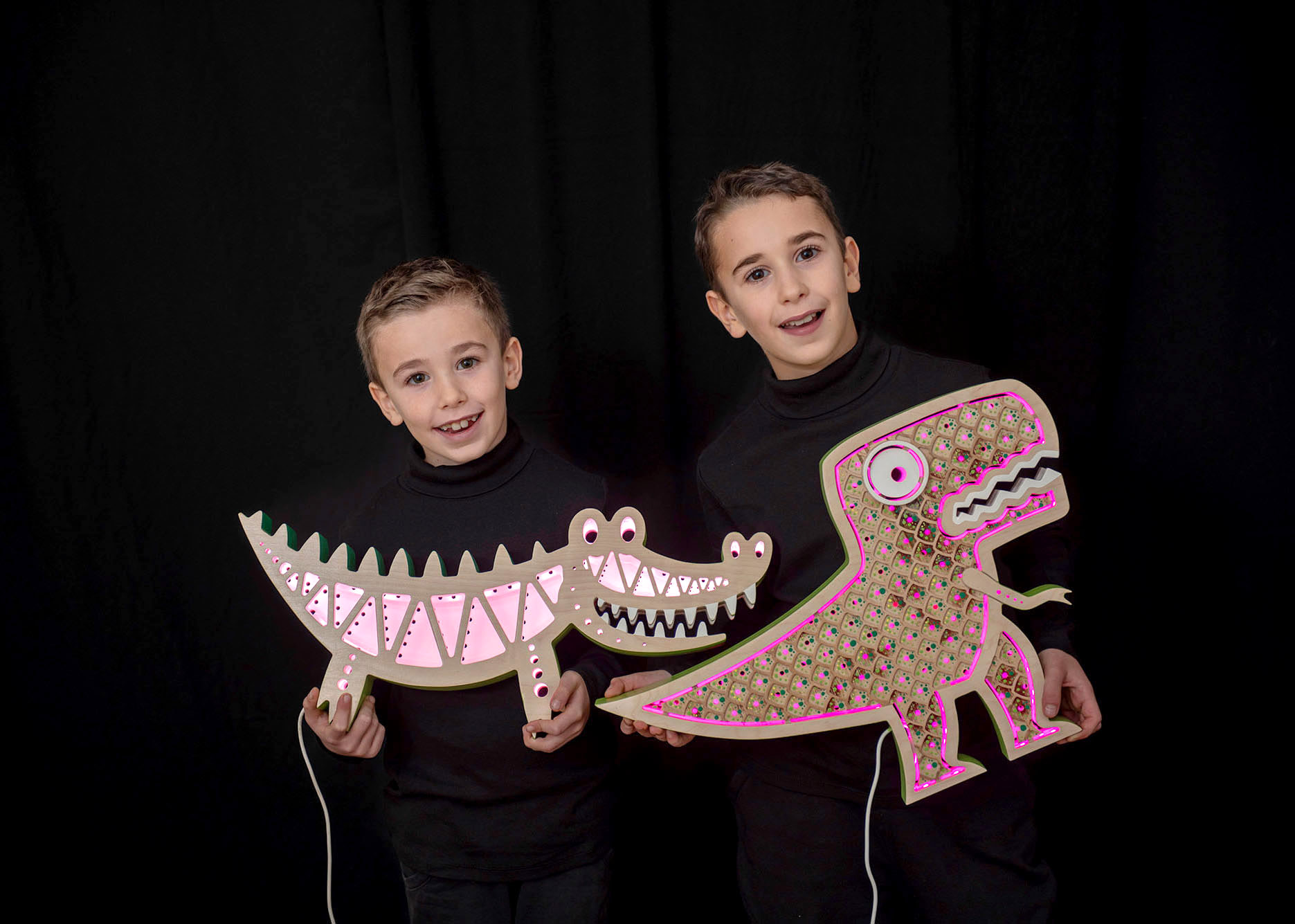 Ritratto professionale di due piccoli artigiani in piedi su fondo nero che tengono in mano due lampade colorate, una a forma di coccodrillo e una a forma di dinosauro.