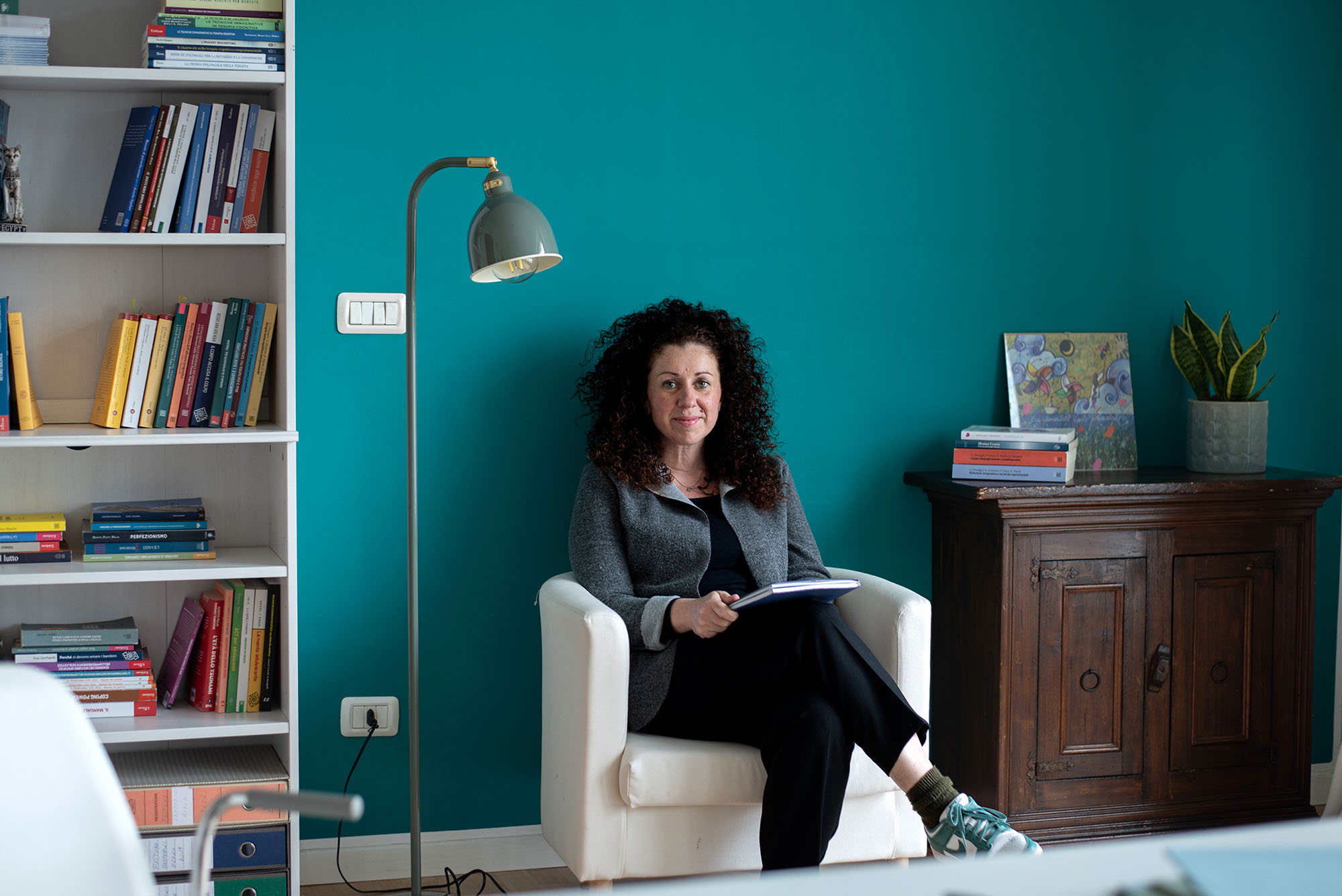 ritratto professionale di una psicologa seduta sulla poltrona del suo studio, davanti a una parete verde con mobile antico e libreria.